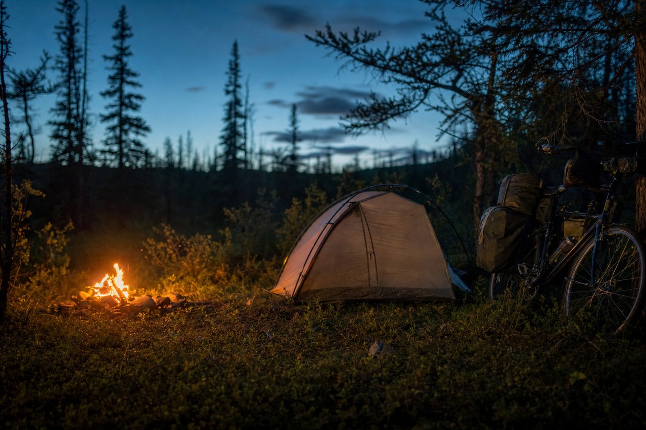 A small tent pitched beside a loaded touring bicycle at dusk beside a campfire