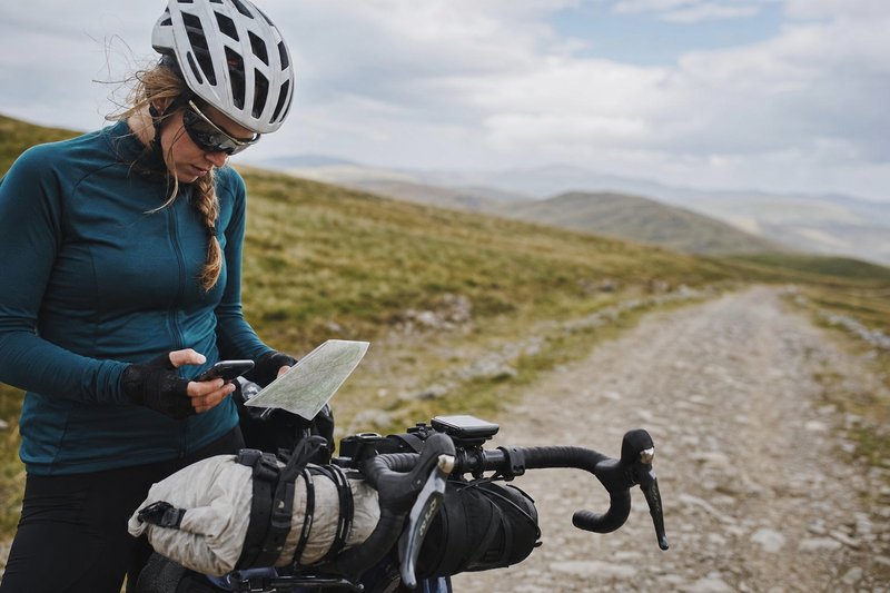 A cyclist stopped on a gravel trail checking a map against handlebar-mounted navigation, loaded bikepacking bike