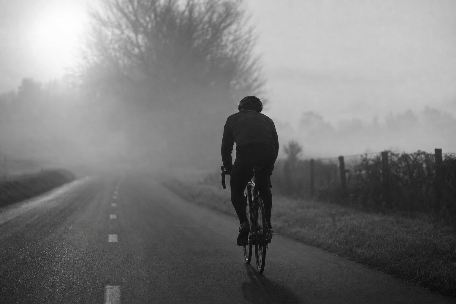 A lone cyclist riding a foggy country road at dawn, black and white silhouette