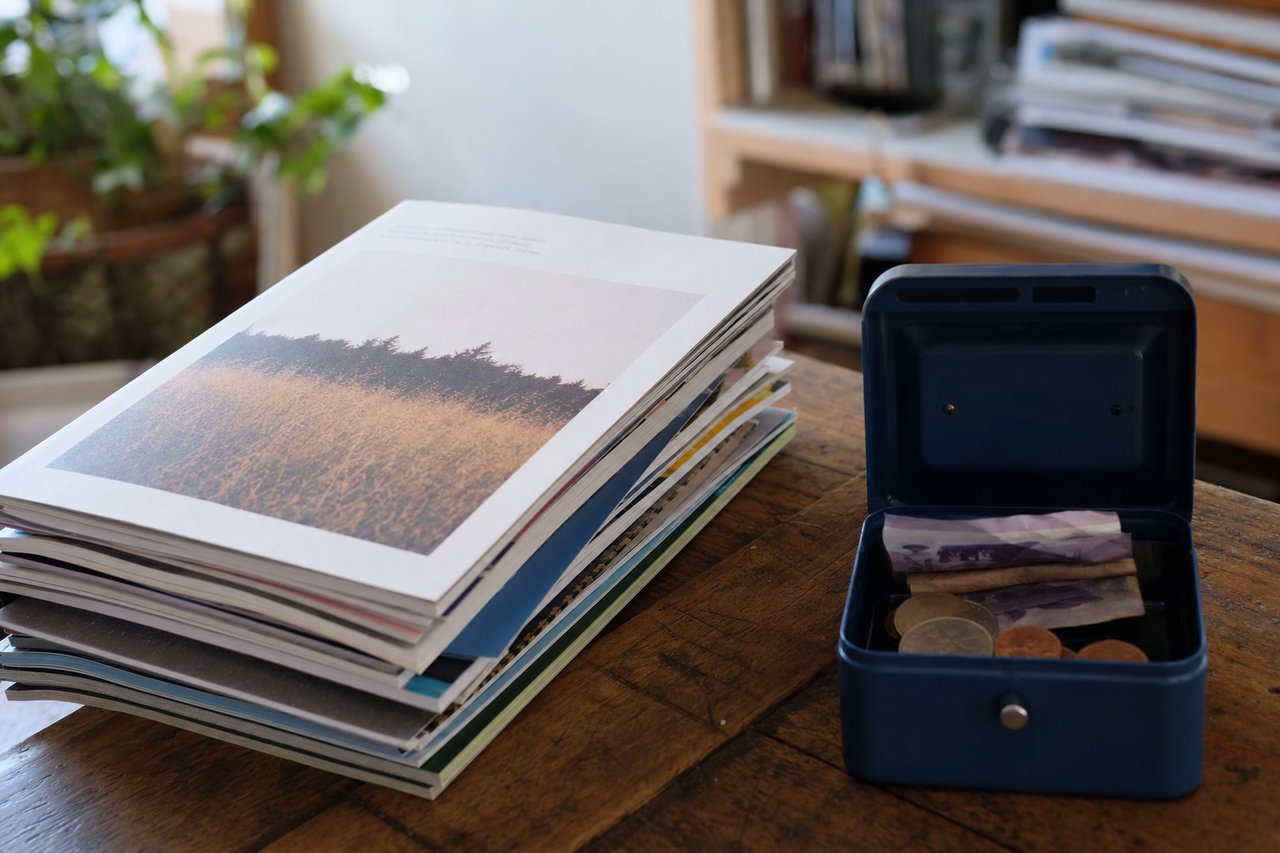 A stack of independent print magazines beside a small cash box of coins and notes, representing the not-for-profit print run